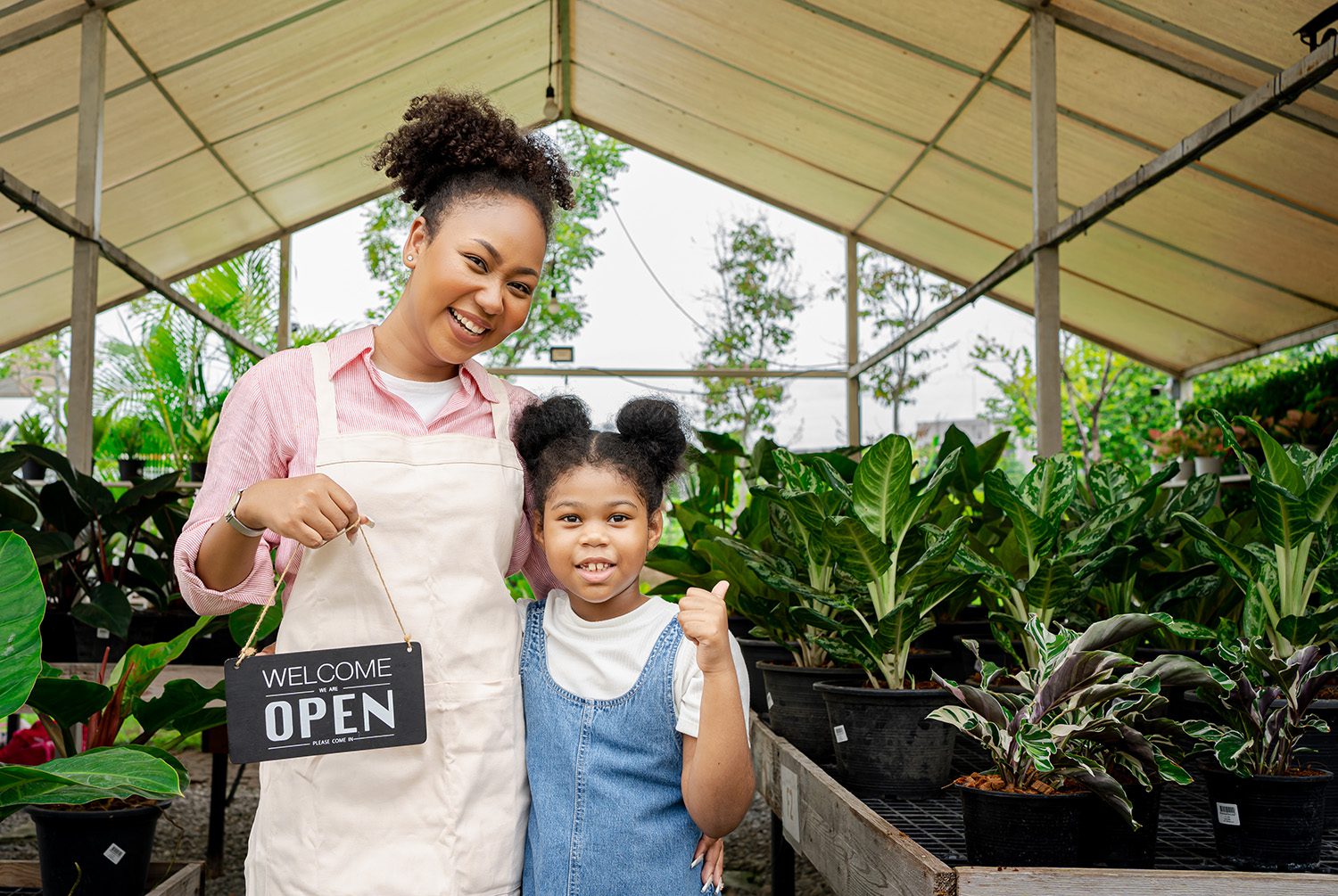 A mom and her daughter smiling at the camera, the mom holding a "Welcome, open" sign and the daughter sporting a thumbs up.