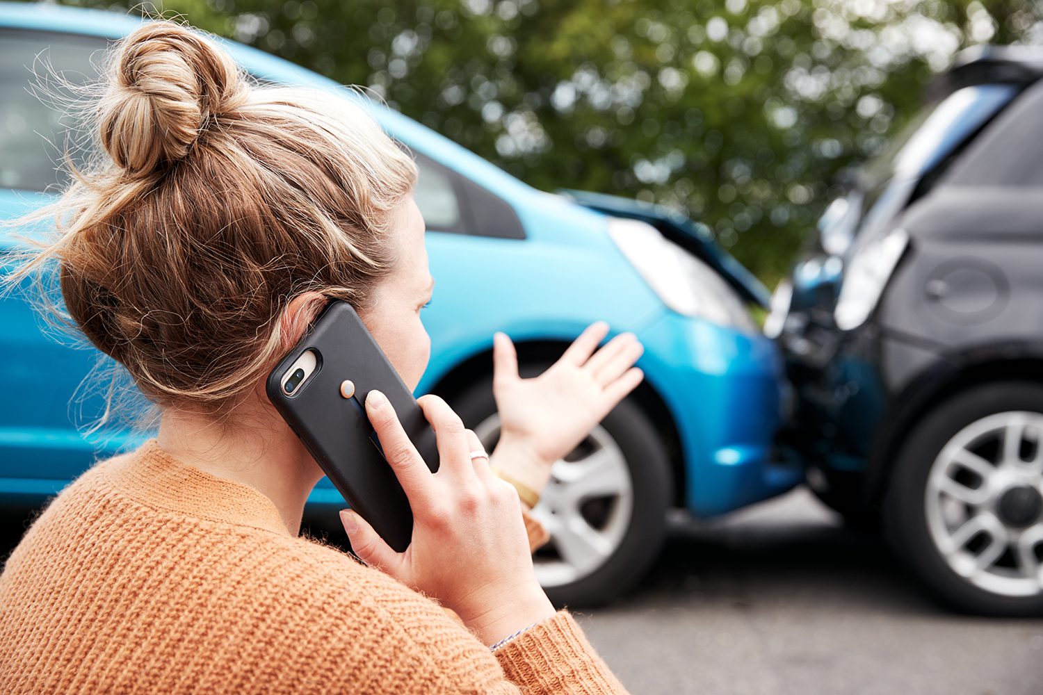 A woman on a phone looking at a car accident and motioning with her hands.
